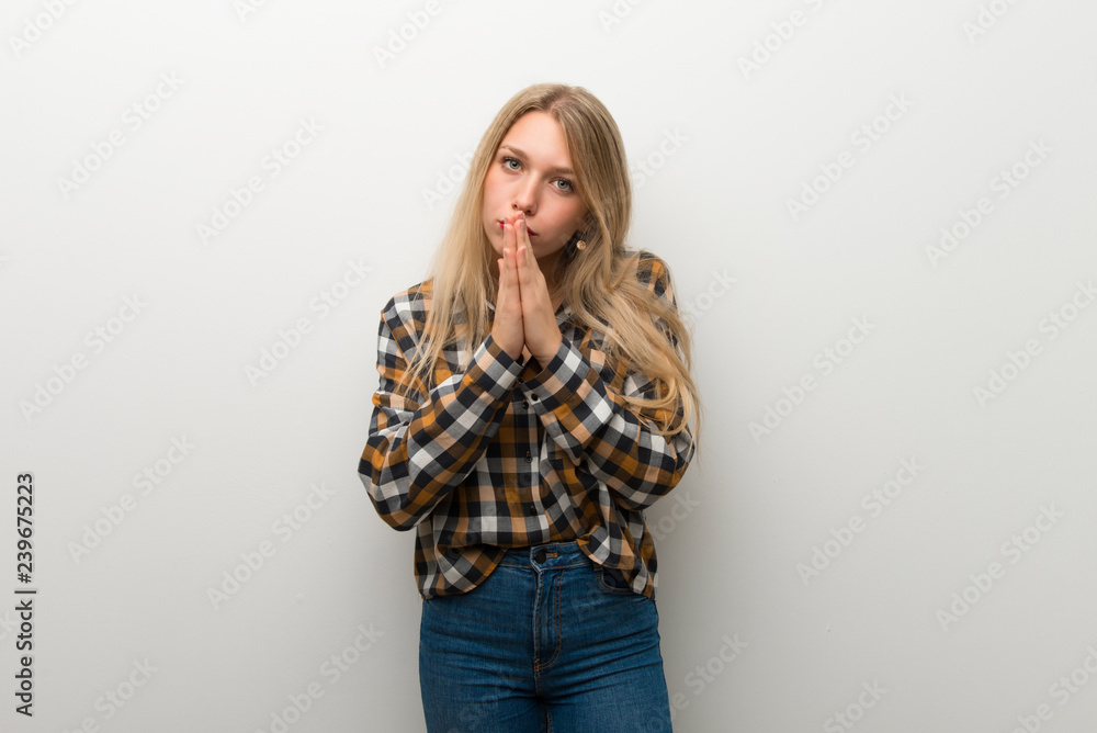 Blonde young girl over white wall keeps palm together. Person asks for something Blonde young girl over white wall keeps palm together. Person asks for something