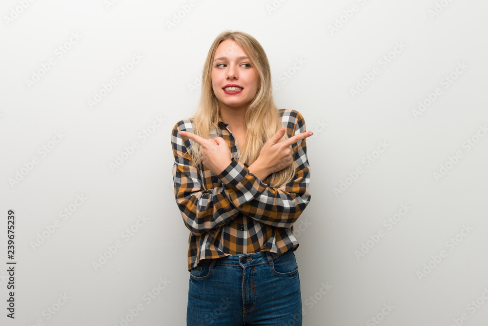 Blonde young girl over white wall pointing to the laterals having doubts Blonde young girl over white wall pointing to the laterals having doubts