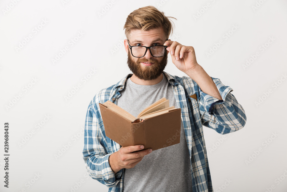 Young bearded man standing isolated over white wall background holding book reading. Young bearded man standing isolated over white wall background holding book reading.