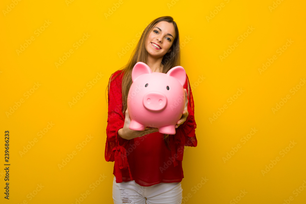 Young girl with red dress over yellow wall holding a piggybank