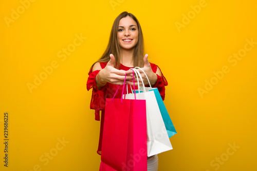 Young girl with red dress over yellow wall holding a lot of shopping bags