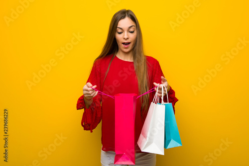 Young girl with red dress over yellow wall surprised while holding a lot of shopping bags