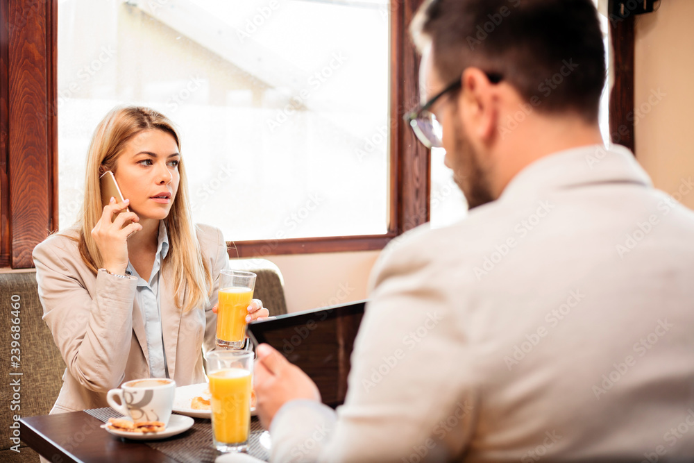 Two young male and female business partners having a meeting over breakfast in a cafe. Woman is talking on the phone and drinking orange juice Two young male and female business partners having a meeting over breakfast in a cafe. Woman is talking on the phone and drinking orange juice