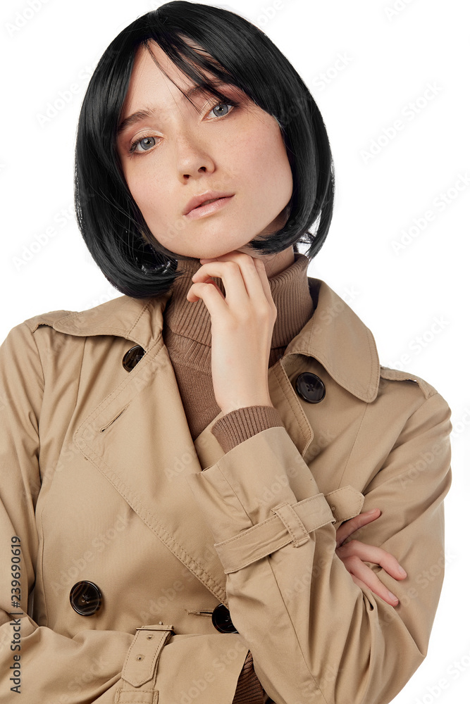 A portrait of a lady, tilting her head and touching her chin. The girl with black short haircut is wearing a trench and sweater. The girl is posing on the white background, looking at the camera.
