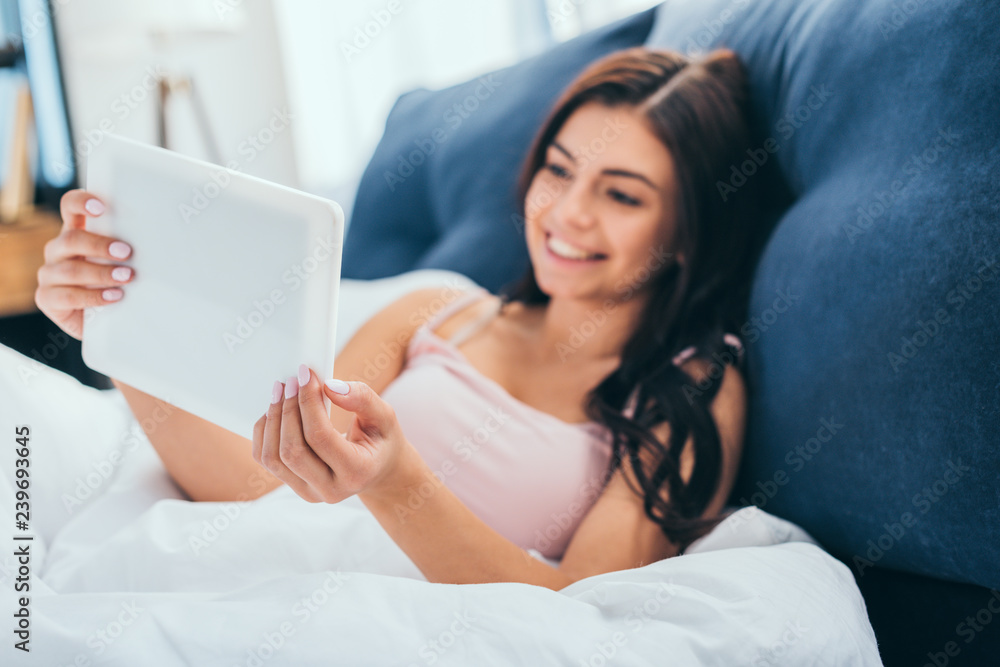 selective focus of smiling woman using digital tablet on bed during morning time at home selective focus of smiling woman using digital tablet on bed during morning time at home