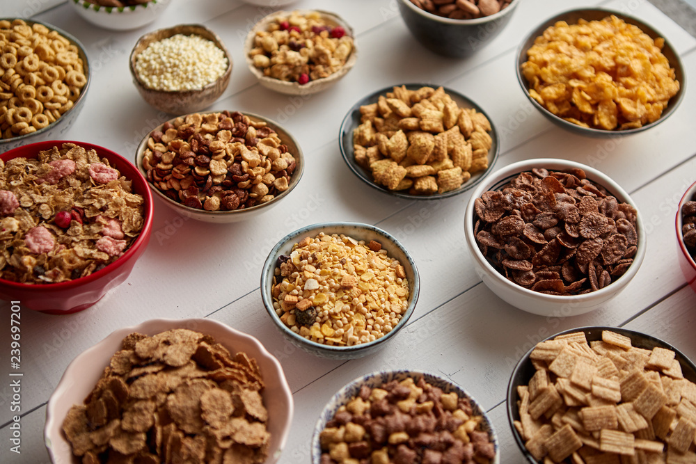 Assortment of different kinds cereals placed in ceramic bowls with cornflakes, granola, cereals and oatmeal. The concept of breakfast food. Flat lay, top view on white wooden table. Assortment of different kinds cereals placed in ceramic bowls with cornflakes, granola, cereals and oatmeal. The concept of breakfast food. Flat lay, top view on white wooden table.