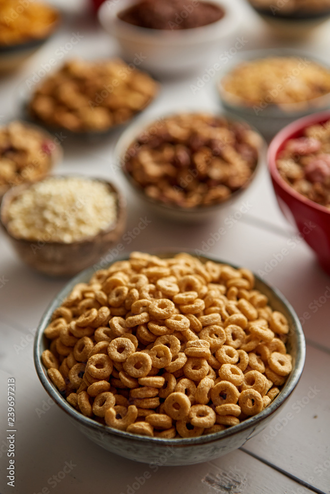 Close up and selective focus. Composition of different kinds cereals placed in ceramic bowls with cornflakes, granola, cereals and oatmeal. Flat lay, top view on white wooden table. Close up and selective focus. Composition of different kinds cereals placed in ceramic bowls with cornflakes, granola, cereals and oatmeal. Flat lay, top view on white wooden table.