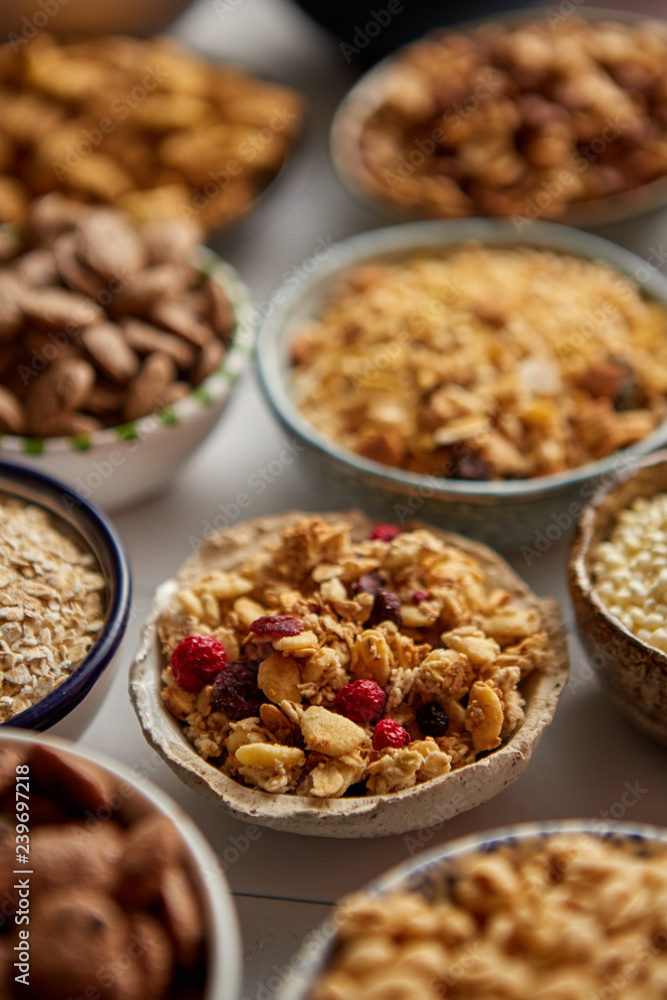 Close up and selective focus. Composition of different kinds cereals placed in ceramic bowls with cornflakes, granola, cereals and oatmeal. Flat lay, top view on white wooden table. Close up and selective focus. Composition of different kinds cereals placed in ceramic bowls with cornflakes, granola, cereals and oatmeal. Flat lay, top view on white wooden table.