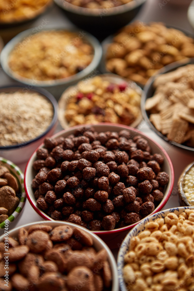Close up and selective focus. Composition of different kinds cereals placed in ceramic bowls with cornflakes, granola, cereals and oatmeal. Flat lay, top view on white wooden table. Close up and selective focus. Composition of different kinds cereals placed in ceramic bowls with cornflakes, granola, cereals and oatmeal. Flat lay, top view on white wooden table.