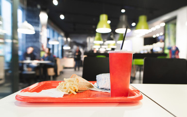 Red tray with Fast Food menu is on the table in the restaurant. French fries, burger, red glass with a cola in the tray against the background of a fast food restaurant. Harmful, caloric food.