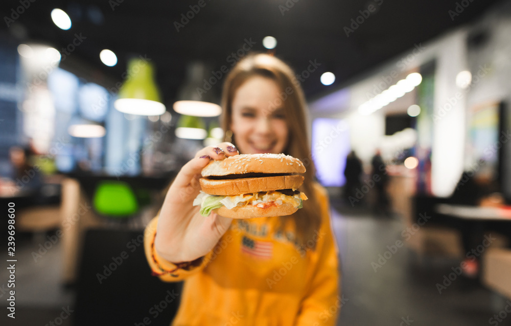 Smiling girl sits at the table in a fast food restaurant and shows an appetizing big burger to the camera.Delicious burger is in the hands of a positive girl. Focus on the burger.