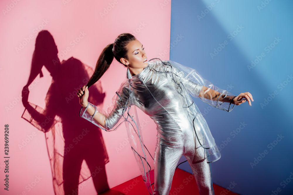 Fototapeta premium beautiful elegant girl posing in metallic bodysuit and trendy raincoat for fashion shoot on pink and blue background