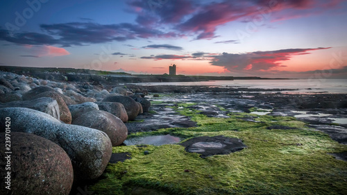Big round rocks and stones with green seaweed at the coast of Easkey Beach, Ireland, with in the background the ruin of a castle-tower under the blue and purple sky after sunset photo