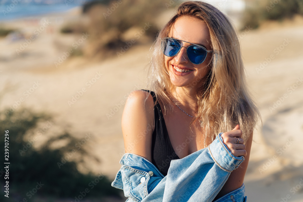 Close-up portrait of beautiful smileing blonde model in black swimsuit and sunglasses posing on the beach. A girl with a sexy figure is holding a denim jacket in her hands, sunbathing on vacation. Close-up portrait of beautiful smileing blonde model in black swimsuit and sunglasses posing on the beach. A girl with a sexy figure is holding a denim jacket in her hands, sunbathing on vacation.