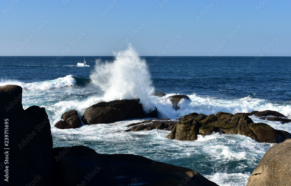 Big waves splashing against the rocks and fishing boat. Blue sea with white foam, sunny day. Galicia, Spain.
