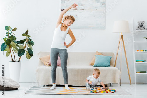 smiling woman doing exercise and looking at cute toddler boy playing with multicolored cubes on carpet in living room