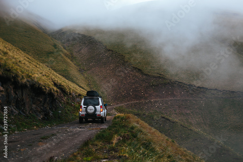 Silver off-road car on road trip in mountains. Compact SUV goes uphill on narrow dangerous road at steep mountainside in the fog. Caucasus Mountains