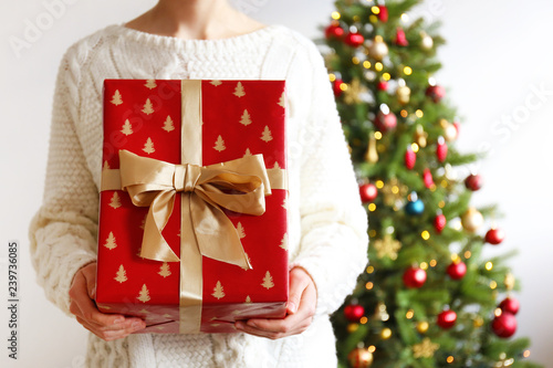 Cropped shot of unrecognizable young woman wearing white knitted sweater, holding a present with festive red wrapping, tied with golden bow, christmas tree on background. Close up, copy space.