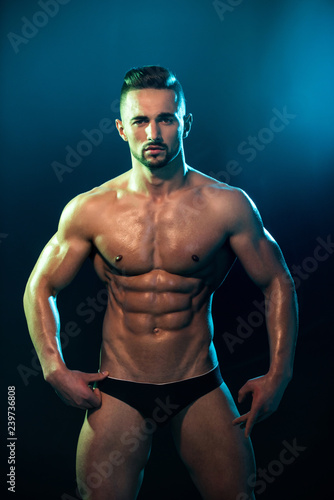 Muscular tanned man in a beautiful pose on a white background. Poledancer on background before night show. Seductive young man and advertising of fitness trainings. Club and entertainment for adults.