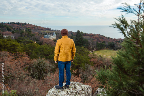 A traveler in a yellow jacket looks down from a mountain on a pine forest. Tourism, active recreation.