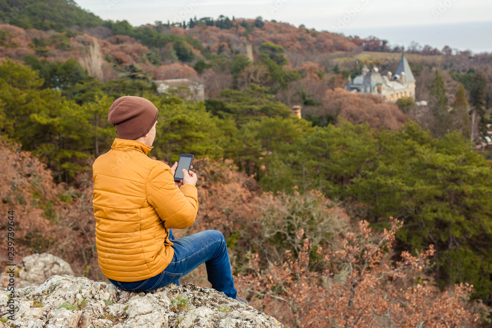 The traveler of the yellow jacket looks with the phone in his hands from the mountain to the pine forest. Tourism, active recreation. The traveler of the yellow jacket looks with the phone in his hands from the mountain to the pine forest. Tourism, active recreation.