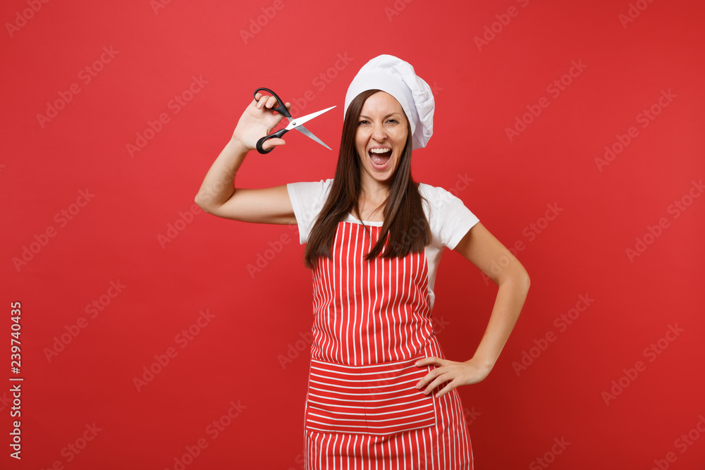 Housewife female chef cook or baker in striped apron, white t-shirt, toque chefs hat isolated on red wall background. Fun crazy housekeeper woman holding kitchen scissors. Mock up copy space concept. Housewife female chef cook or baker in striped apron, white t-shirt, toque chefs hat isolated on red wall background. Fun crazy housekeeper woman holding kitchen scissors. Mock up copy space concept.