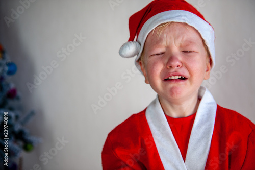 Funny child in Santa Claus costume is crying without Christmas present. Tradition of family holidays.