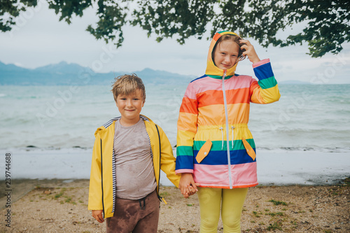 Group of two funny kids playing by the lake on a very windy day, watching waves, wearing rain coats. Image taken on lake Geneva, Lausanne, Switzerland