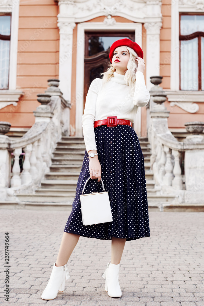 hellige knus Krydret Foto Stock Outdoor full body fashion portrait of young beautiful girl  wearing red beret, belt, turtleneck, polka dot midi skirt, white ankle boots,  heels, holding small bag, posing in old european street 