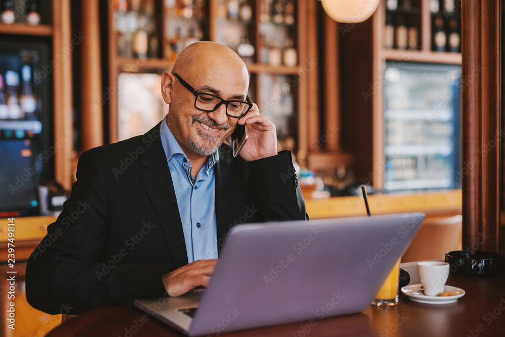 Bearded senior in suit using smart phone and laptop while sitting in cafeteria. On the table laptop, juice and coffee. Bearded senior in suit using smart phone and laptop while sitting in cafeteria. On the table laptop, juice and coffee.