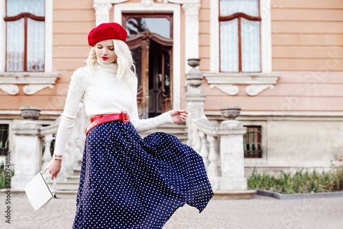 Outdoor fashion portrait of young beautiful woman wearing red beret, belt, turtleneck, polka dot blue midi skirt, holding small white leather bag, posing in old european street. Copy, empty space 