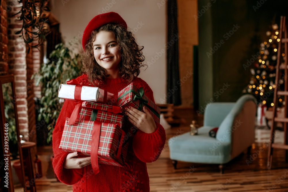 Christmas, New Year, winter holidays concept: young happy smiling lady holding a lot of packaged gifts, posing at festive decorated home. Model wearing red sweater, beret. Copy, empty space for text