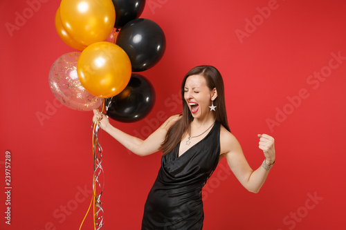 Overjoyed young woman in black dress celebrating screaming, doing winner gesture, holding air balloons isolated on red background. Valentine's Day Happy New Year birthday mockup holiday party concept. © ViDi Studio