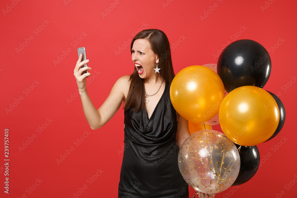 Fototapeta premium Angry young woman in little black dress holding air balloons, making video call, talking on mobile phone, screaming isolated on red background. Happy New Year, birthday mockup holiday party concept.