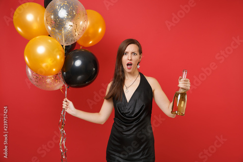 Shocked bewildered young woman in black dress holding bottle of champagne, air balloons isolated on red background. International Women's Day, Happy New Year, birthday mockup holiday party concept.