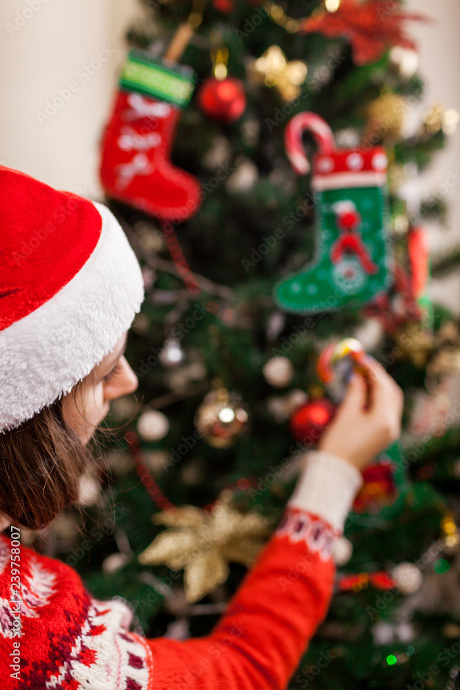 Young woman decorating Christmas tree with candy canes at home, wearing Santa hat. Preparing to New year.