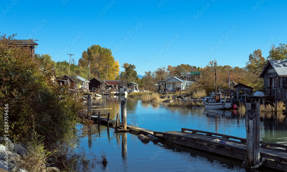 An old fishermen village located in the slough of river. Old houses with moorings and boats near the shore. Early autumn, old grass and yellow trees against the blue sky. 