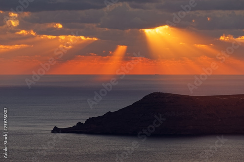 Moody sunset with orange-red light on the sea  clouds in front of the sun create rays of light and shadow  in the foreground the silhouette of an island - Greece  Cyclades  Santorini  Santorin  Thira 