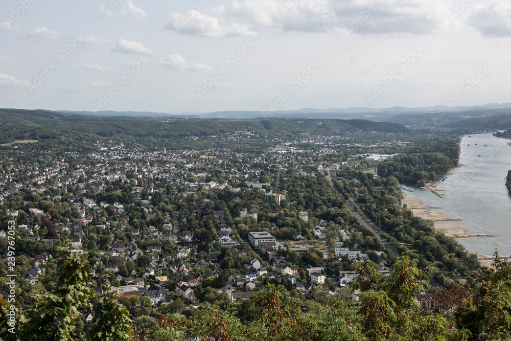 Bird's eye view of a city from Drachenfels Hills. The city looks congested with lush greeneries and mountains. A river can be seen flowing with boats floating on it.
