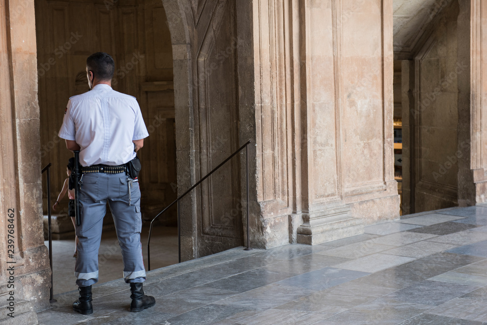 Alhambra Palace security guard keeping watch during the day. The cop is seen wearing uniform of white shirt and grey pants. He is leaning against the wall.