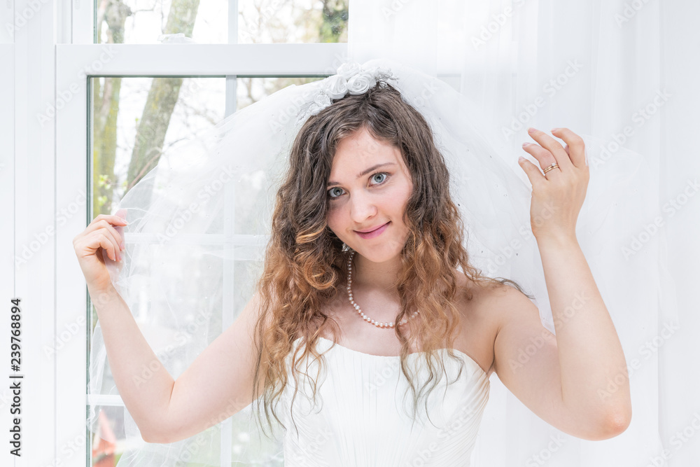 Closeup portrait of young female person, woman, bride in wedding dress, holding veil, hands, face, pearl necklace, standing by window, white curtains, happy, smiling in engagement ring Closeup portrait of young female person, woman, bride in wedding dress, holding veil, hands, face, pearl necklace, standing by window, white curtains, happy, smiling in engagement ring