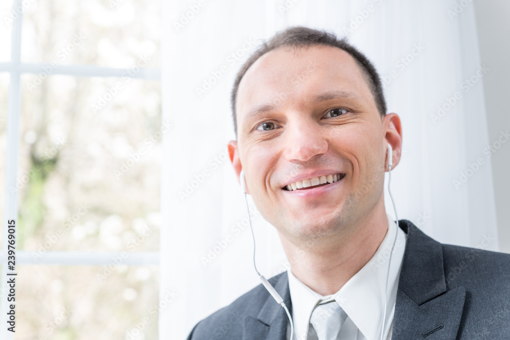 Closeup of young, happy smiling man, male businessman in business suit, tie, standing by window in home, house room, white curtains, with earbuds headphones, talking, listening to music Closeup of young, happy smiling man, male businessman in business suit, tie, standing by window in home, house room, white curtains, with earbuds headphones, talking, listening to music