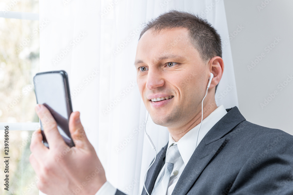 Closeup of young, happy man, male businessman in business suit, tie, standing by window in home, house room, white curtains, with earbuds headphones, holding phone, talking, video conferencing Closeup of young, happy man, male businessman in business suit, tie, standing by window in home, house room, white curtains, with earbuds headphones, holding phone, talking, video conferencing