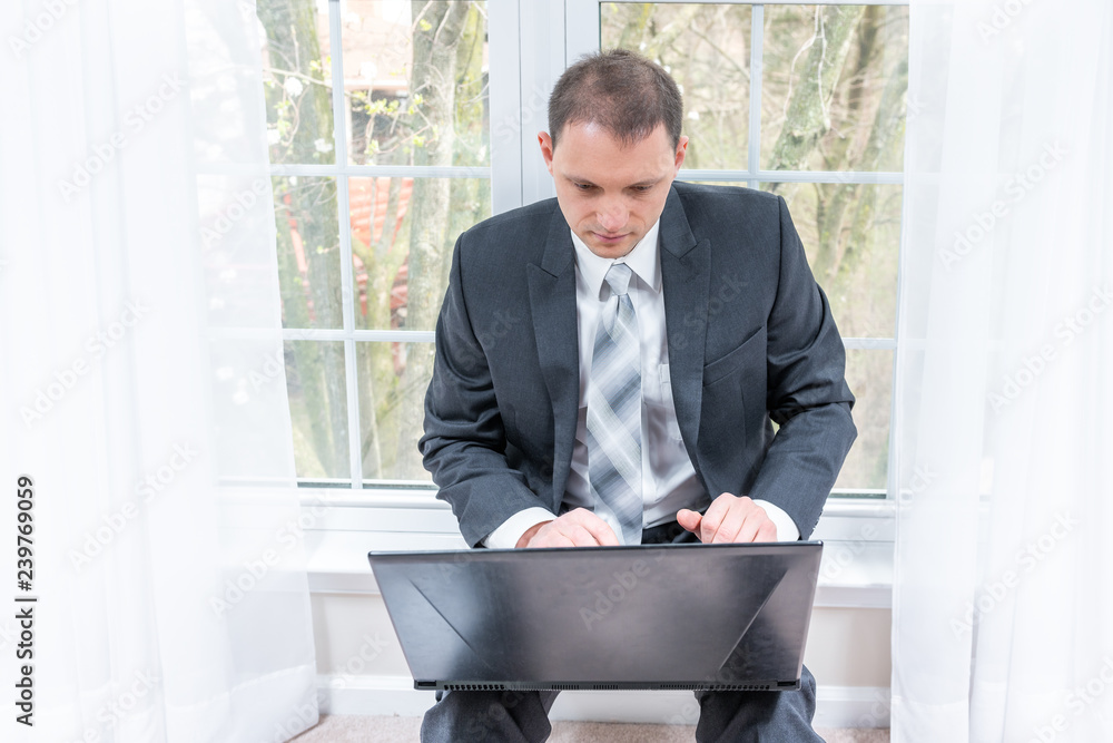 Young man, businessman, business suit, tie, sitting in house, home, apartment room on windowsill, window sill with white curtains, working on black laptop computer, notebook on lap, legs Young man, businessman, business suit, tie, sitting in house, home, apartment room on windowsill, window sill with white curtains, working on black laptop computer, notebook on lap, legs