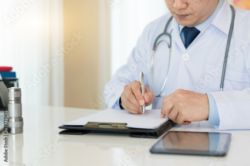 Doctor writing on medical health care record, patients discharge, or prescription form paperwork in hospital clinic office with physician's stethoscope on neck..