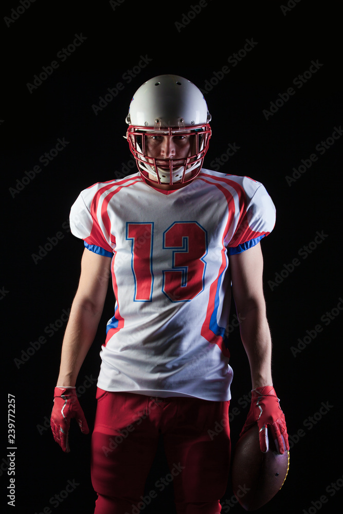 American football player wearing helmet posing with ball on black background