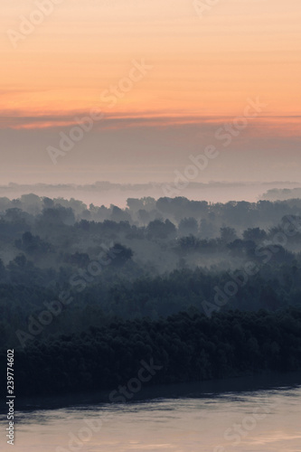 Mystical view on riverbank of large island with forest under haze at early morning. Eerie mist among layers from tree silhouettes. Morning atmospheric landscape of majestic nature in blue faded tones.