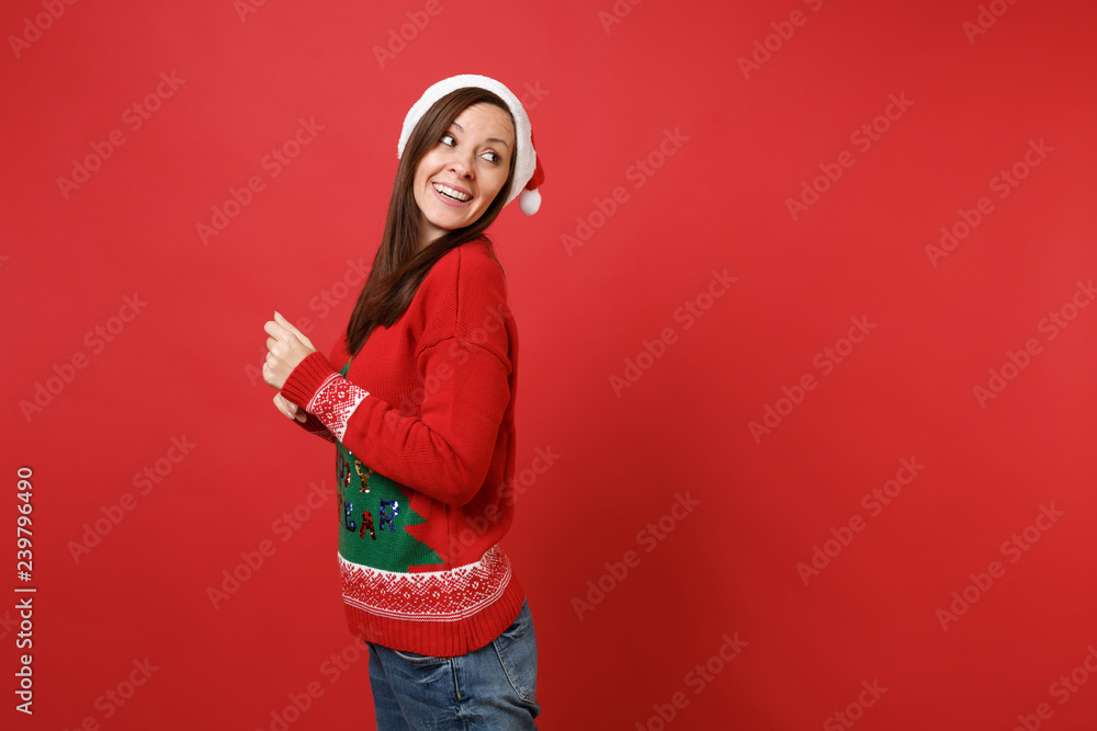 Side view of pretty young Santa girl in knitted sweater, Christmas hat looking back isolated on bright red wall background. Happy New Year 2019 celebration holiday party concept. Mock up copy space. Side view of pretty young Santa girl in knitted sweater, Christmas hat looking back isolated on bright red wall background. Happy New Year 2019 celebration holiday party concept. Mock up copy space.