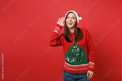 Curious young Santa girl in knitted sweater, Christmas hat eavesdrop, hearing gesture isolated on bright red wall background. Happy New Year 2019 celebration holiday party concept. Mock up copy space.