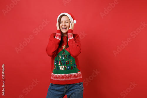 Laughing young Santa girl with closed eyes in Christmas hat putting hands on cheeks, head isolated on bright red background. Happy New Year 2019 celebration holiday party concept. Mock up copy space.